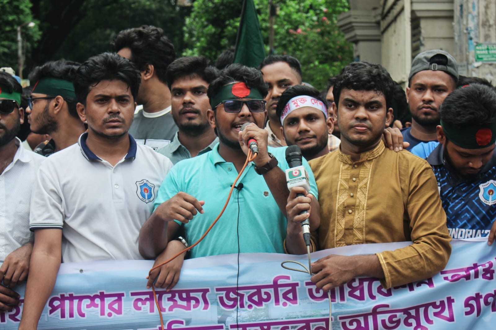 মাথায় বাংলাদেশের পতাকার ব্যান্ড পরে ব্যানারের সামনে মাইকে বক্তব্য